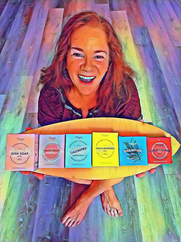 A bright, multi colored image of a smiling Angie Ringler holding a board with a row of bar soaps on the board